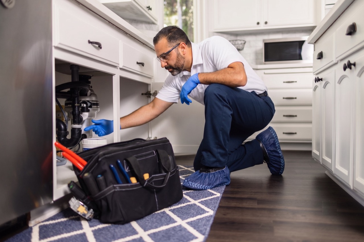 Plumbing technician inspecting pipes under a kitchen sink with a tool bag